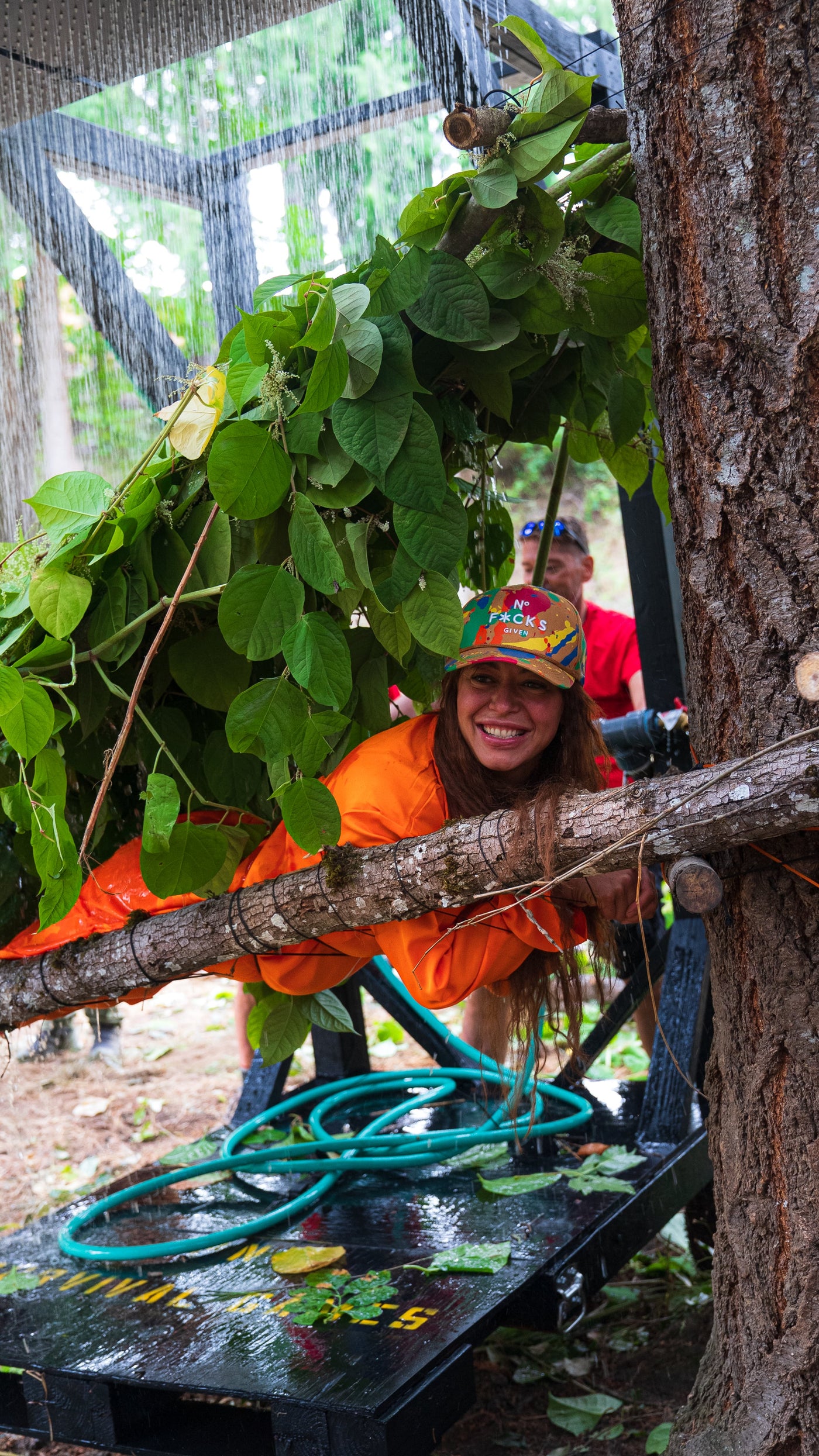 A competitor smiling from a leafy shelter as water pours down during a 2026 Survival Games challenge
